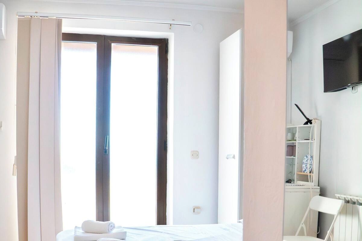A bedroom with a bed, a chair, a TV, and a window.