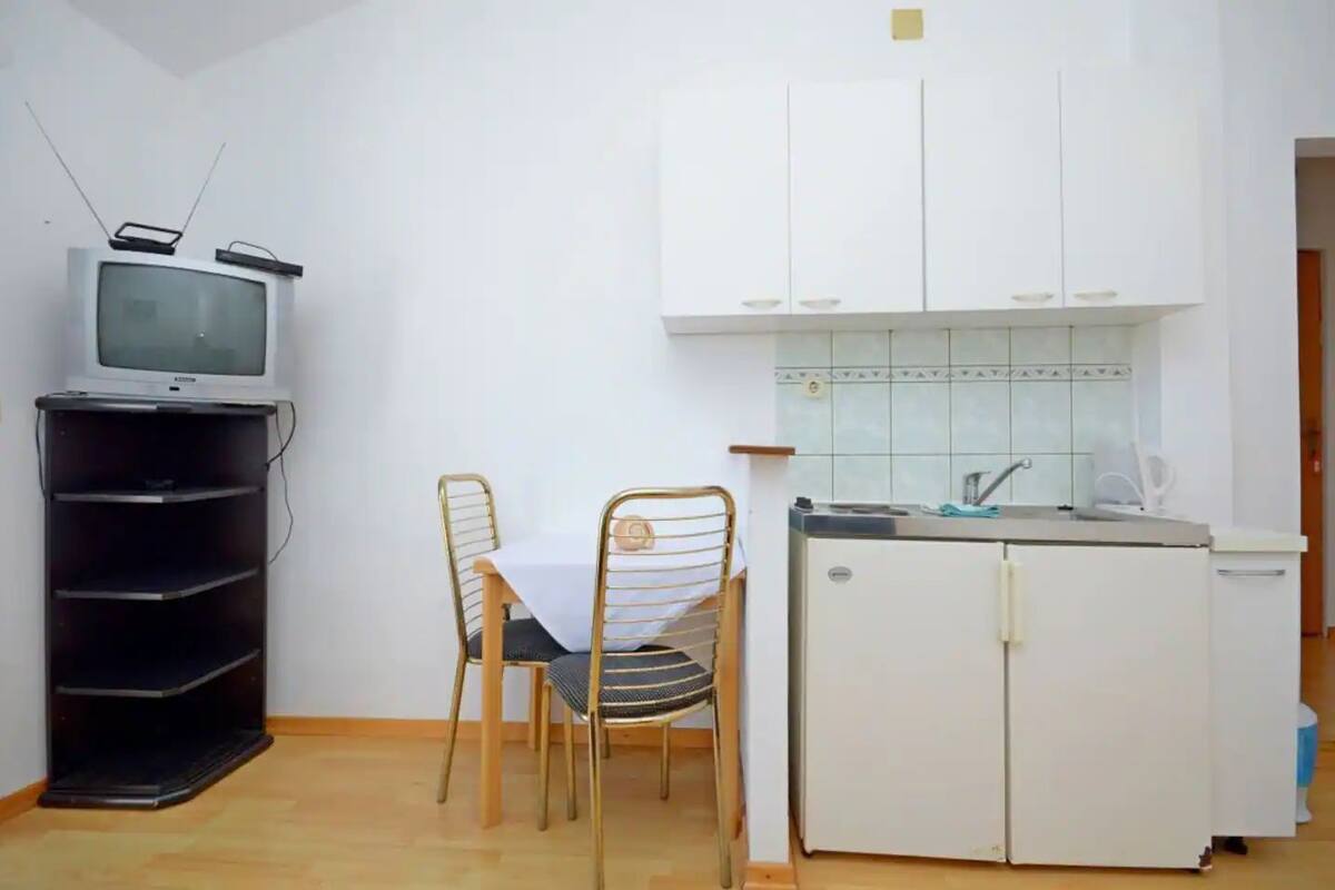 A small, compact kitchen with a refrigerator, sink, and a television on a stand. There is a dining table with two chairs.