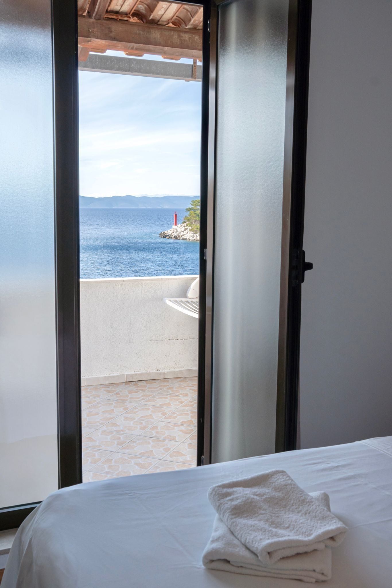 A hotel room with a bed, a white towel, and a view of the sea through an open door.