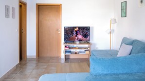 Uma sala de estar com um sofá azul, uma porta de madeira, uma televisão sobre um suporte e um abajur.