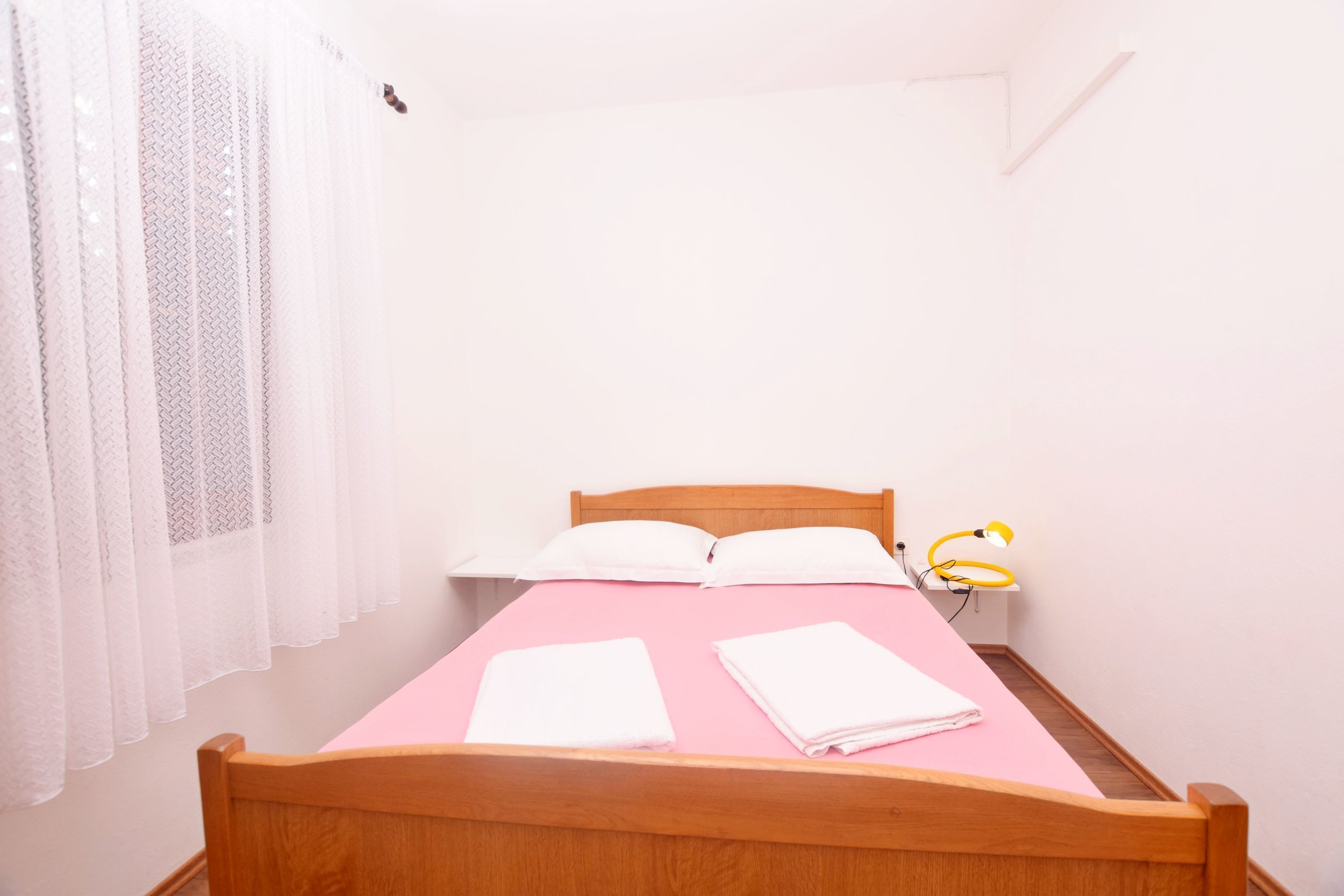 Uma cama de solteiro com estrutura de madeira, colcha rosa, duas almofadas brancas e duas toalhas brancas dobradas.