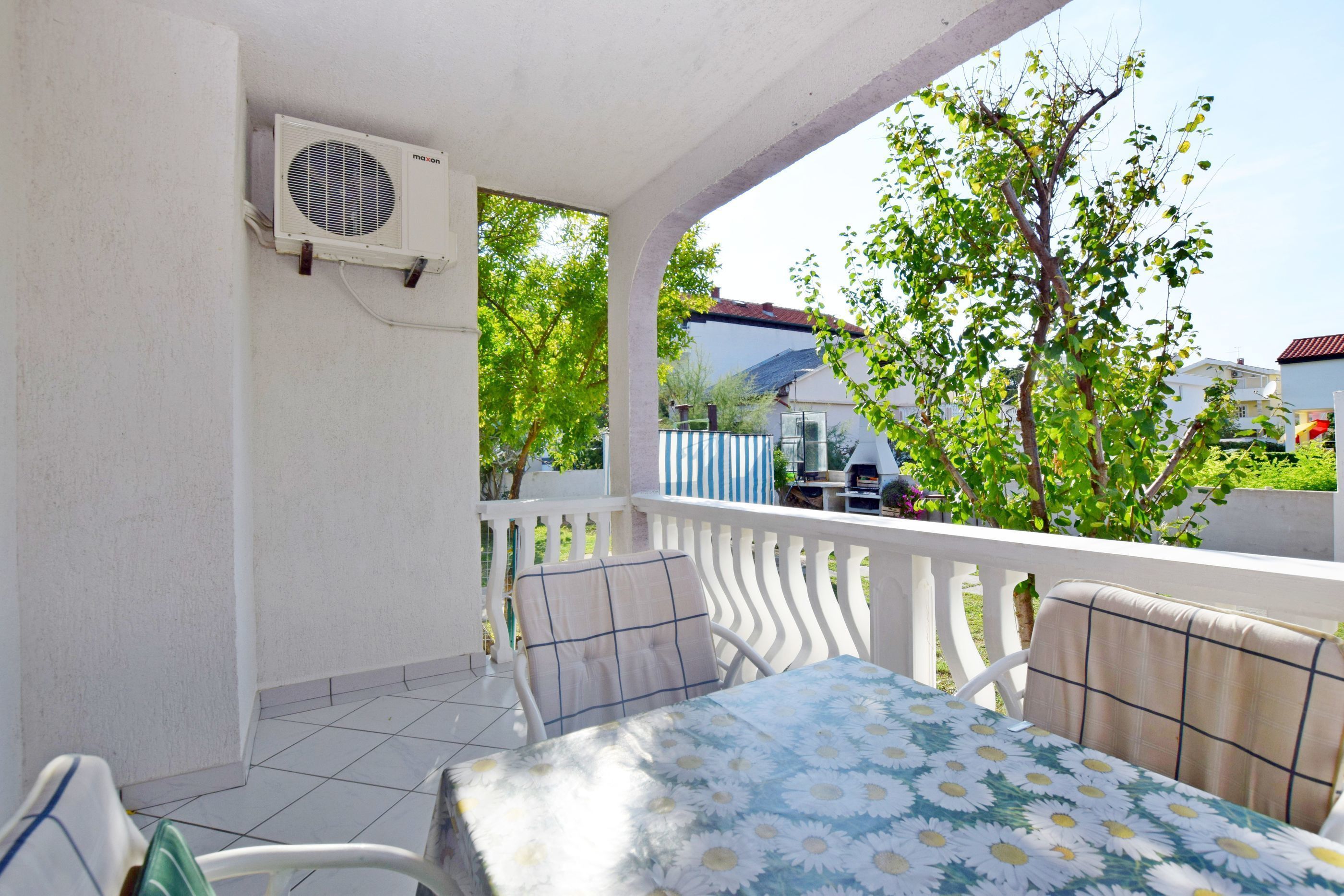 Un balcon avec une table et des chaises, un appareil de climatisation et une vue sur un arbre et une maison au loin.