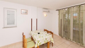 Un coin repas avec une table recouverte d’une nappe à motifs floraux, des chaises en bois, une fenêtre avec des stores et un rideau sur le balcon.
