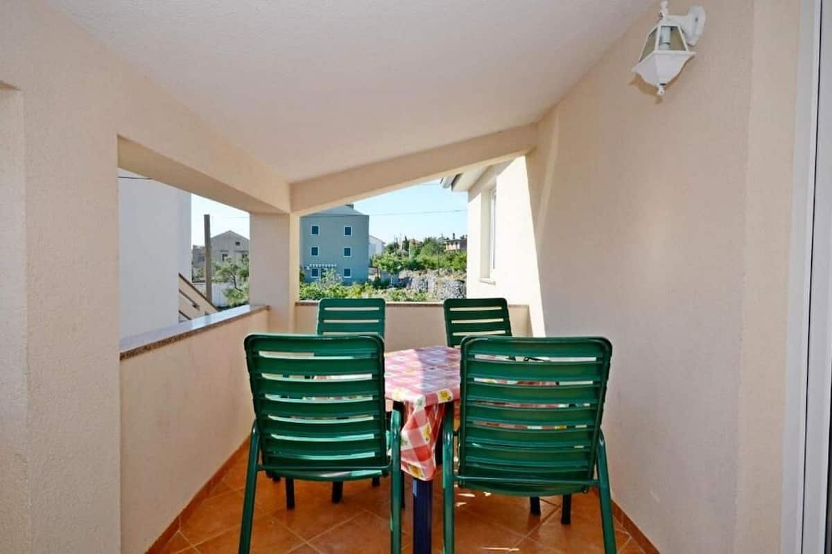 A balcony with a table and four chairs, a view of buildings and greenery, and a white wall with a window.