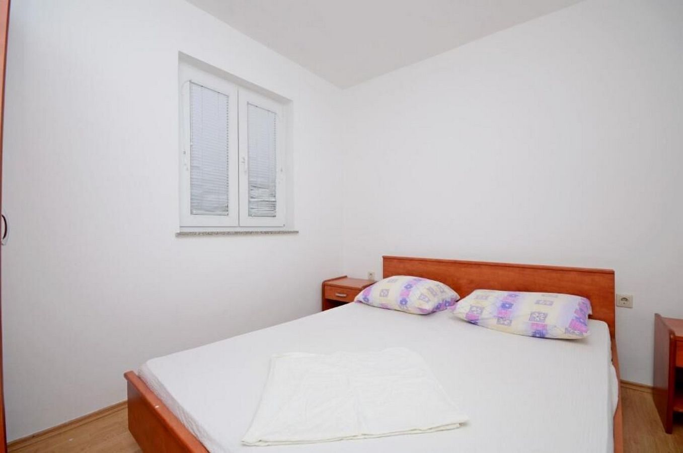 A single bed with a wooden headboard, two pillows, a bedside table, and a window with closed blinds.