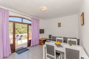 Een kamer met balkon, een eettafel met stoelen, een bank, een aan de muur bevestigde televisie en een raam met gordijnen.
