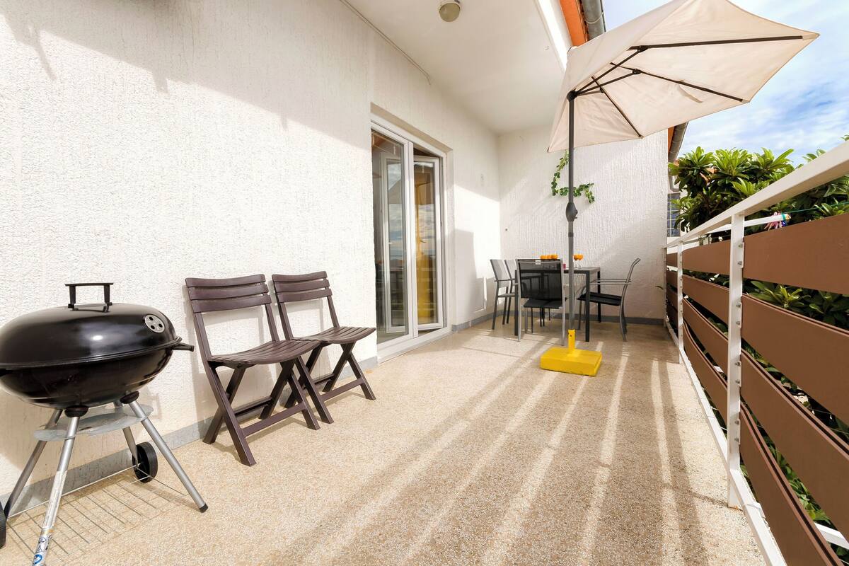 A patio with a barbecue grill, two wooden chairs, a table with chairs, and a large umbrella.