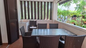 A dining area with a table and chairs, a window with vertical blinds, and a view of greenery outside. - Apartment Jolanka (Sukosan)