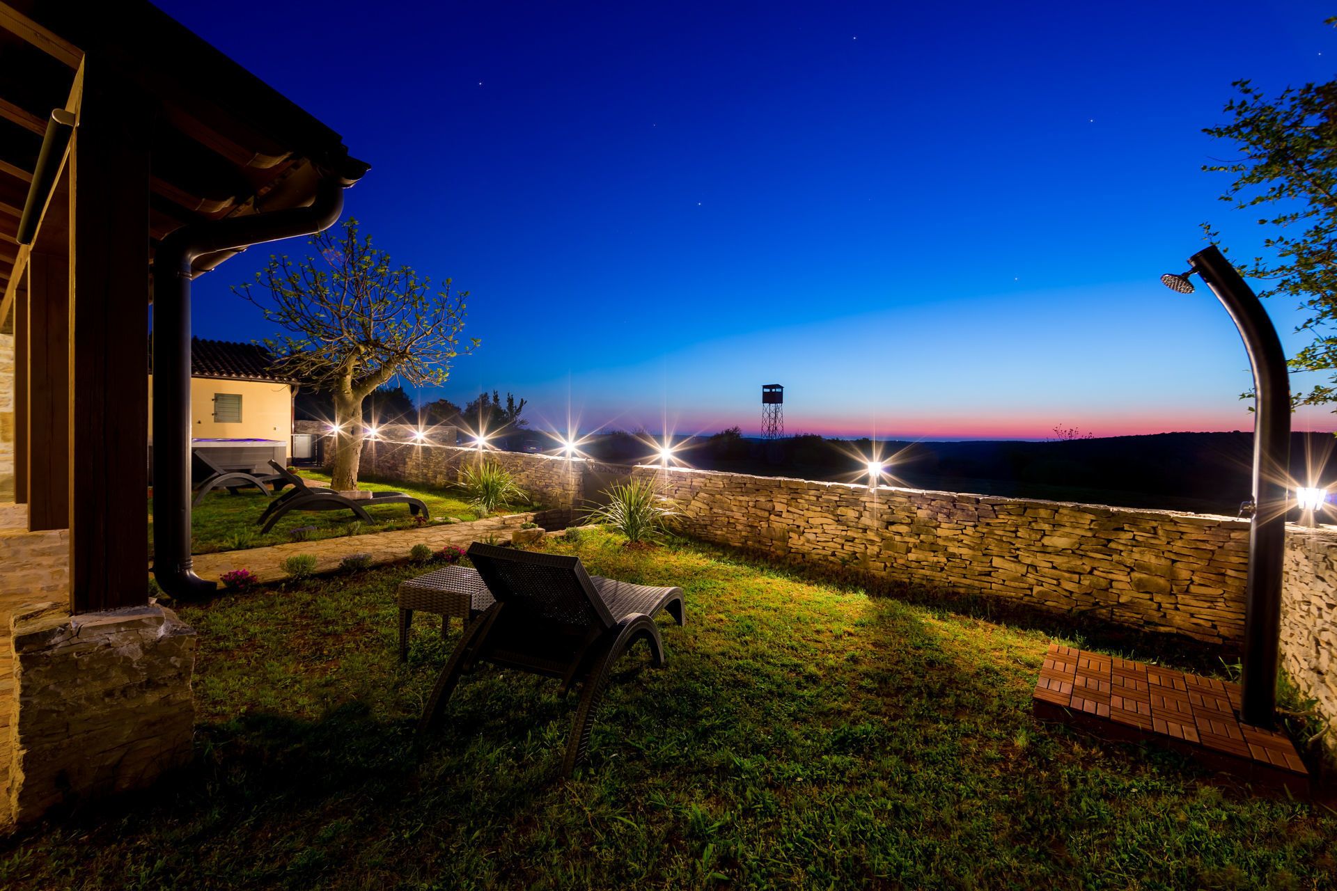 Un'area patio all'aperto con un muro in pietra, una pergola in legno e vista su una torre d'acqua in lontananza.