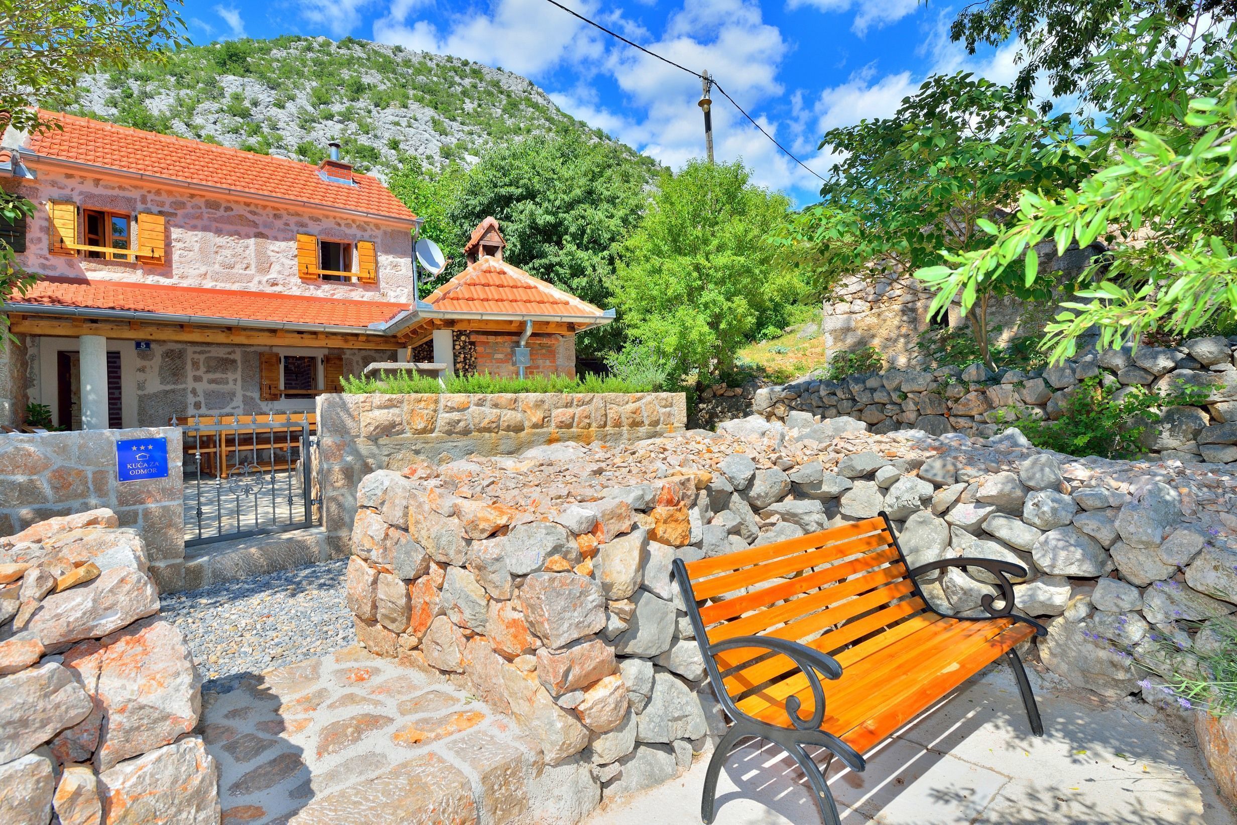 A stone-walled courtyard with a wooden bench, a stone building with a red-tiled roof, and a mountain backdrop.