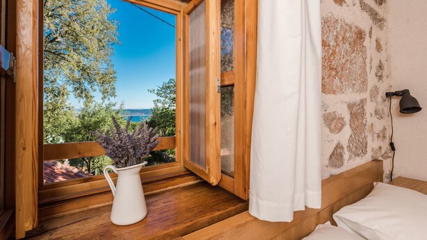 A wooden-framed window with a white curtain, a white vase with flowers, and a view of trees and a body of water. - House Martelina (Starigrad - Paklenica)