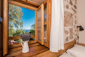 A wooden-framed window with a white curtain, a white vase with flowers, and a view of trees and a body of water. - House Martelina (Starigrad - Paklenica)