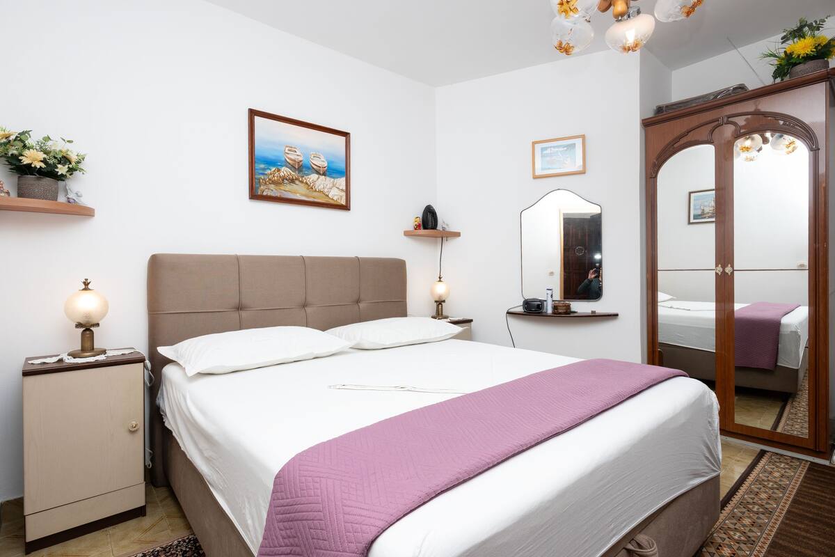 A bedroom with a bed, bedside tables, a wardrobe, a mirror, and wall-mounted shelves with flowers.
