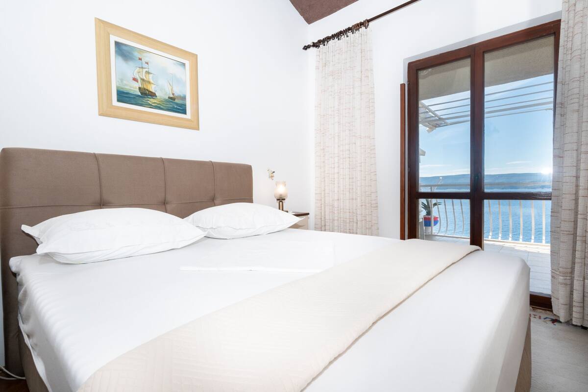 A bedroom with a large bed, a nightstand with a lamp, a framed picture on the wall, and a view of the sea through a window.