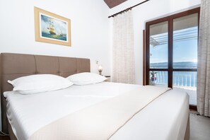 A bedroom with a large bed, a nightstand with a lamp, a framed picture on the wall, and a view of the sea through a window. - Apartments Neno (Stanici)