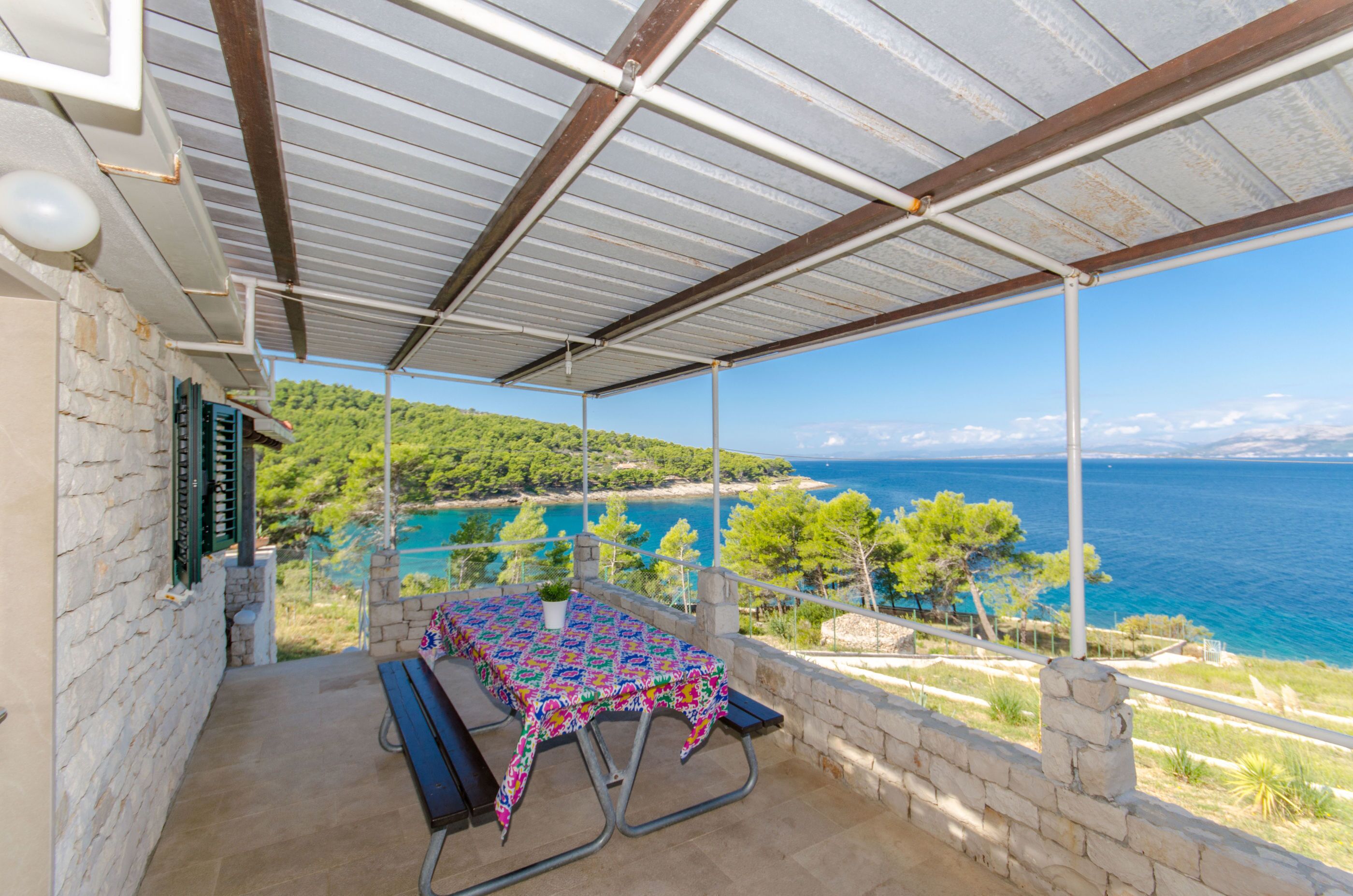 A covered outdoor area with a table and benches, overlooking a coastal landscape with trees and the sea.