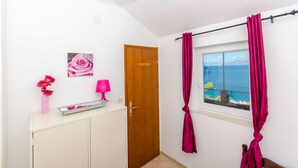 A room with a wooden door, a window with curtains, a small cabinet, a pink vase with flowers, and a pink lamp. - House Romantica (Postira)