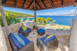 A covered outdoor seating area with wicker furniture and cushions, overlooking a scenic coastal view. - House Romantica (Postira)