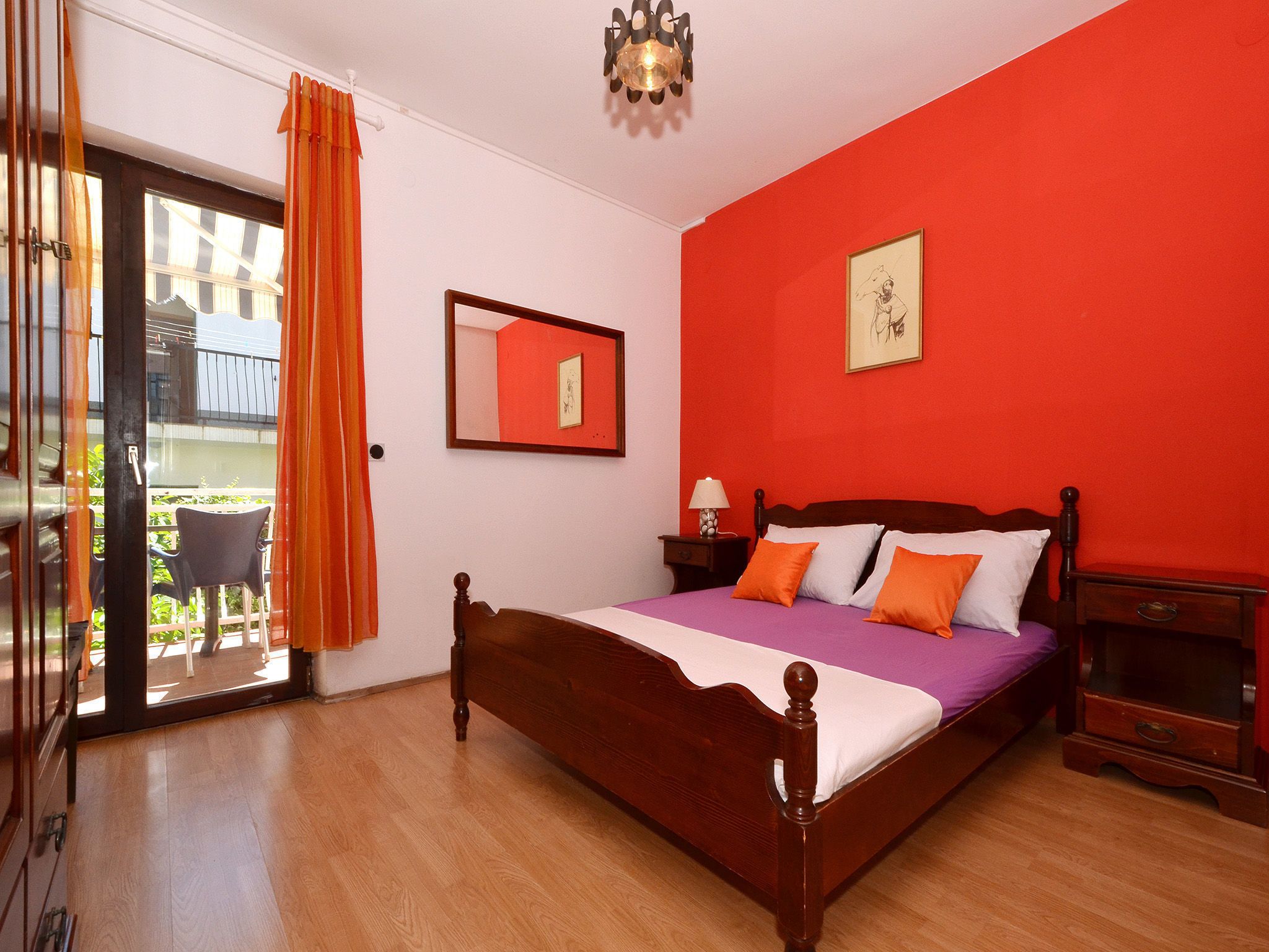 A bedroom with a wooden bed, a red wall, a window with curtains, a small table with a lamp, and a framed picture.