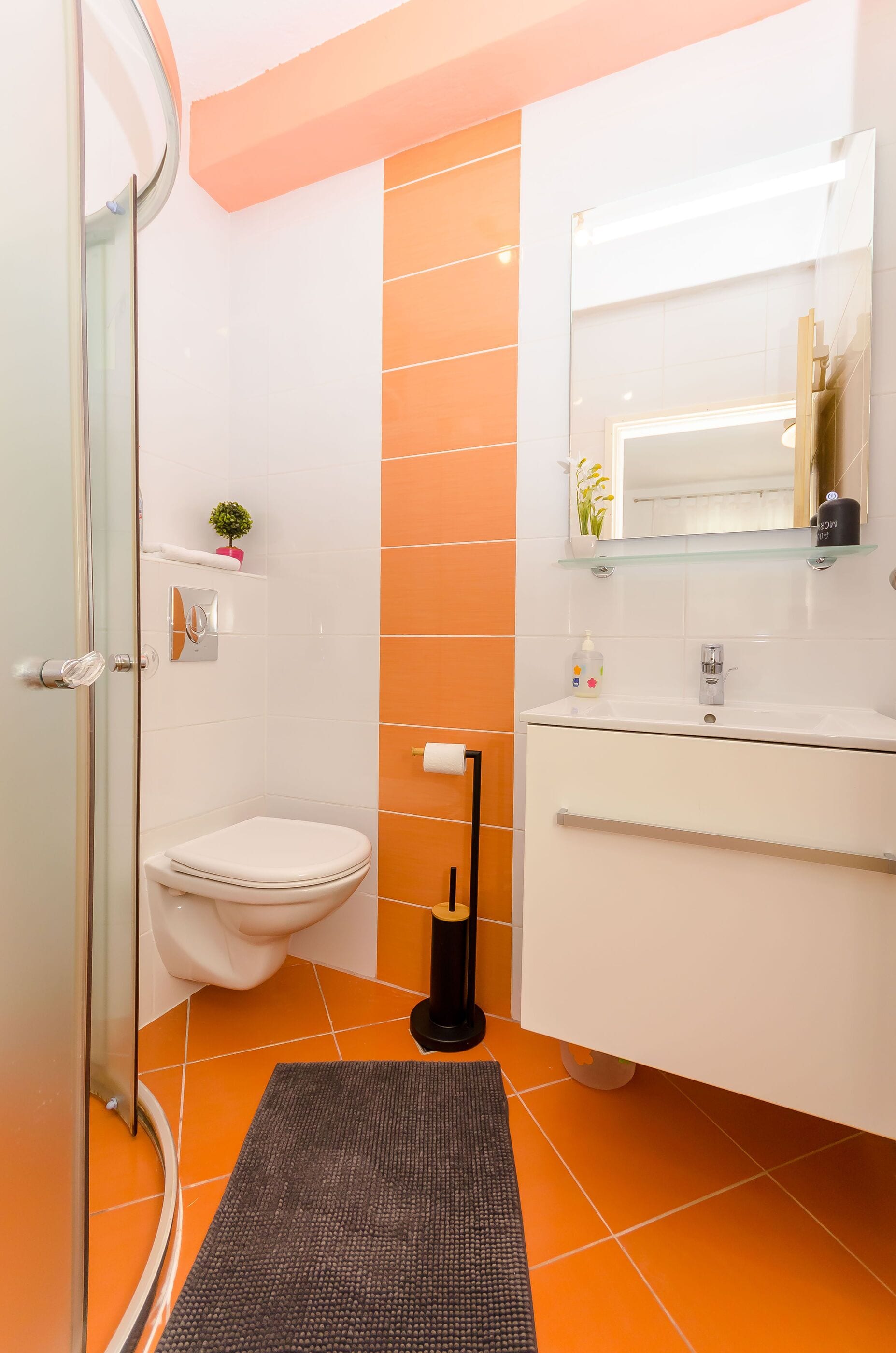 A bathroom with a glass shower enclosure, a white toilet, an orange-tiled wall, a white sink with a mirror above it, and a small potted plant on the shelf.
