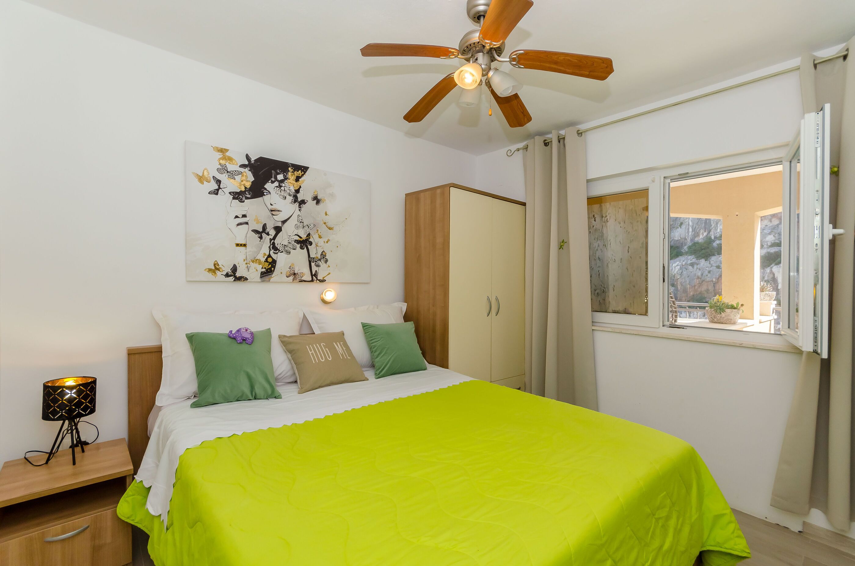 Ein Schlafzimmer mit Bett, Deckenventilator, Nachttisch, Lampe, Kleiderschrank und einem Fenster mit Blick ins Freie.