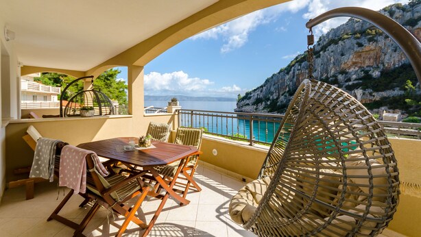 A balcony with a wooden table and chairs, a wicker chair, and a view of the sea and mountains. - Apartments Apollo (Zastrazisce)