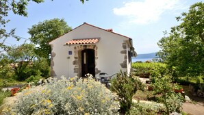 Un petit bâtiment en pierre, surmonté d’un toit de tuiles, entouré d’une végétation luxuriante et de plantes en fleurs.