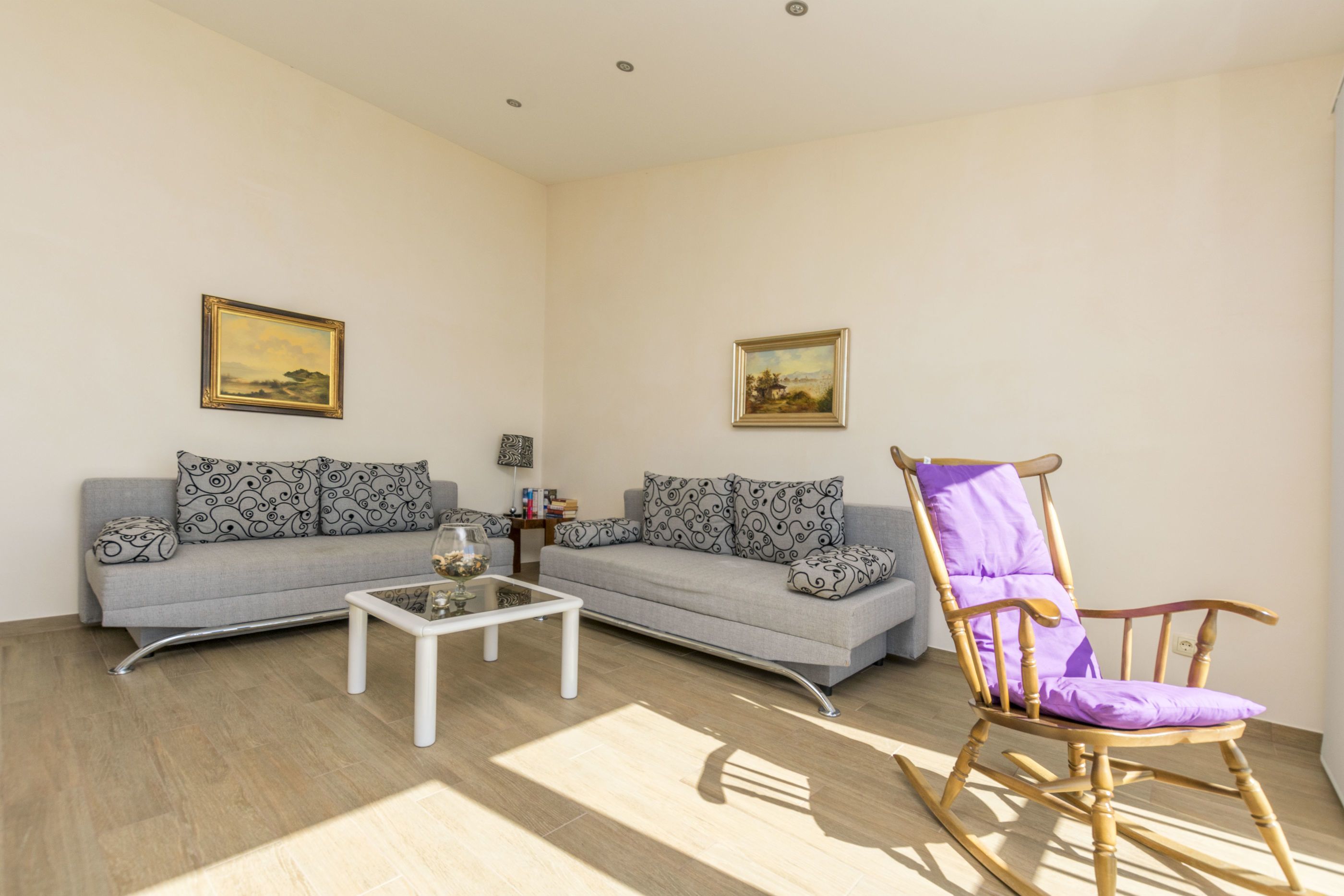 Una sala de estar con un sofá gris, una mesa de centro blanca, un sillón mecedor de madera y dos cuadros enmarcados en las paredes.