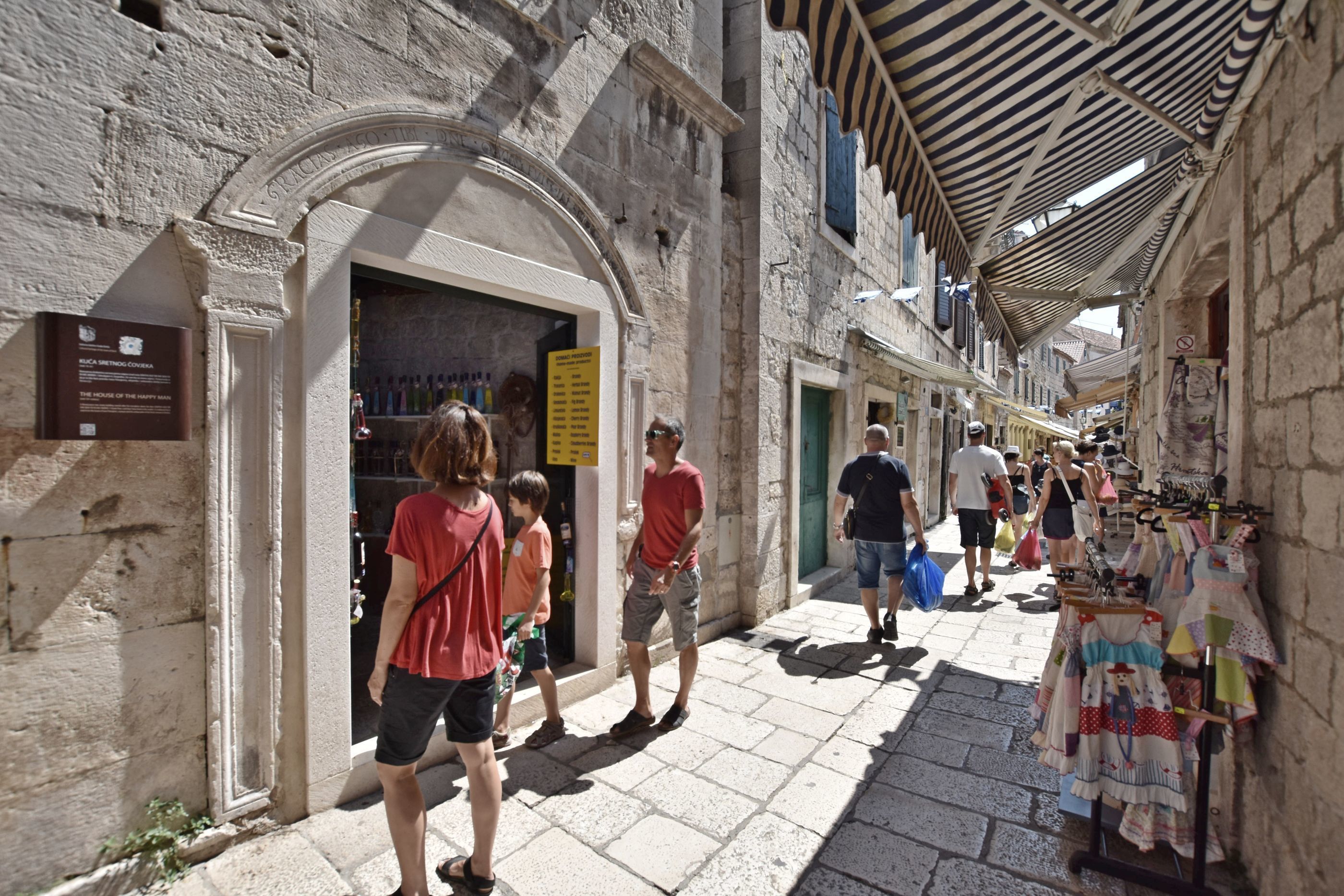 A narrow street with stone buildings, a shop with a signboard, and a clothing stall.