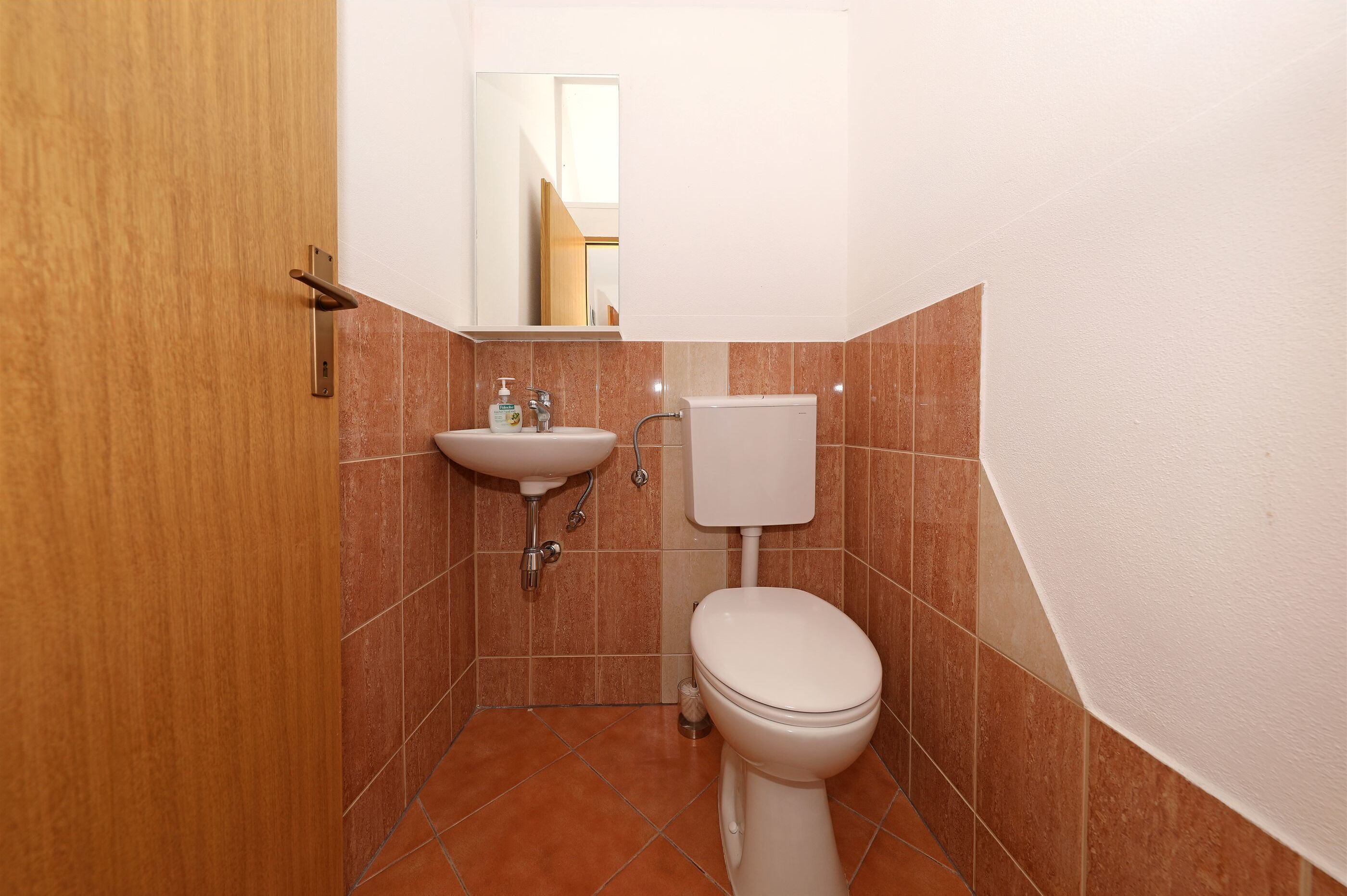Ein kleines Badezimmer mit Holztür, Waschbecken, Toilette und Spiegel.