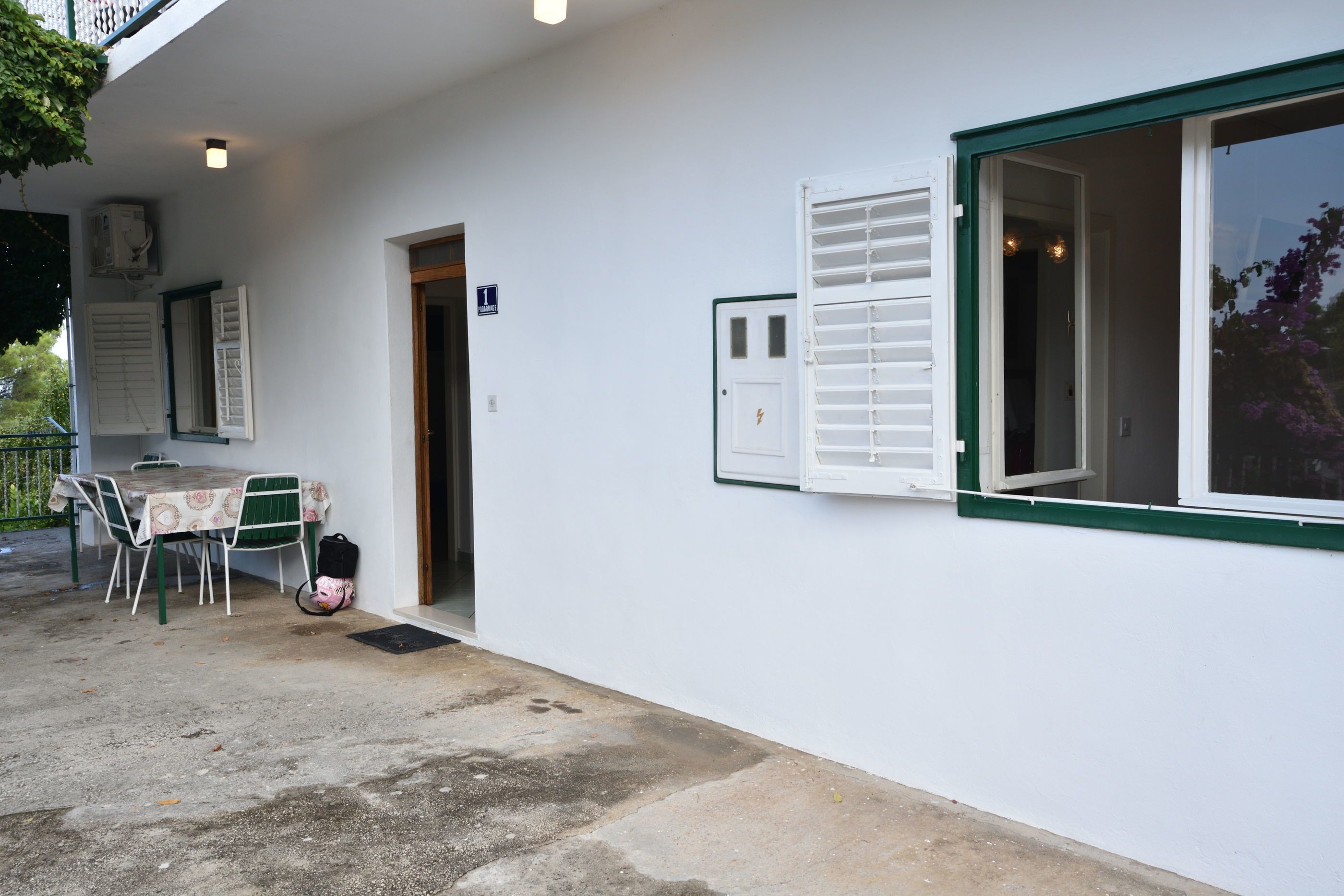 Fachada limpia y blanca de un edificio con un área de estar al aire libre cubierta, una puerta con un cartel y una ventana con contraventanas.