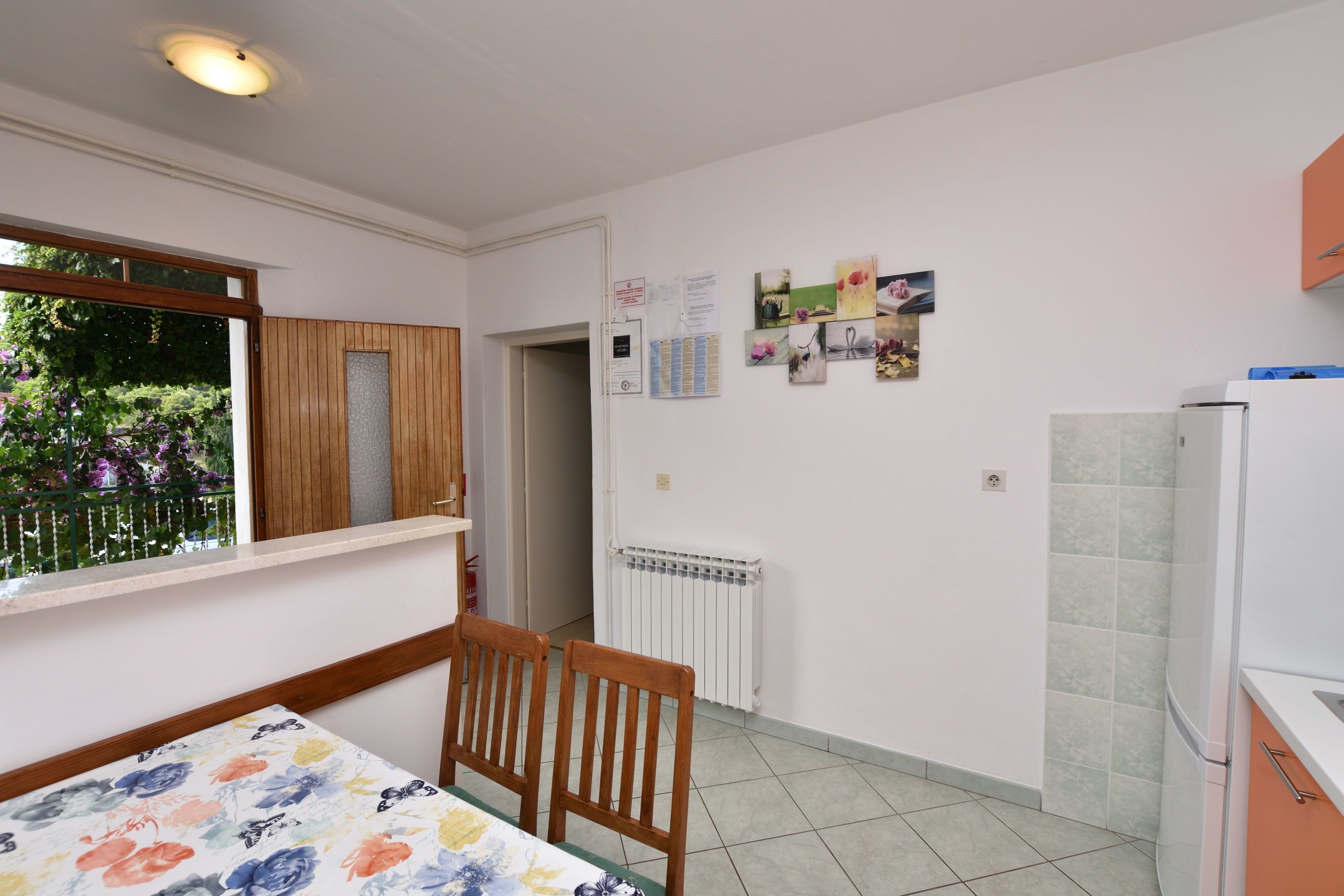 Een kleine, schone kamer met een eettafel, twee stoelen, een raam met uitzicht op groen, een witte radiator en een koelkast.