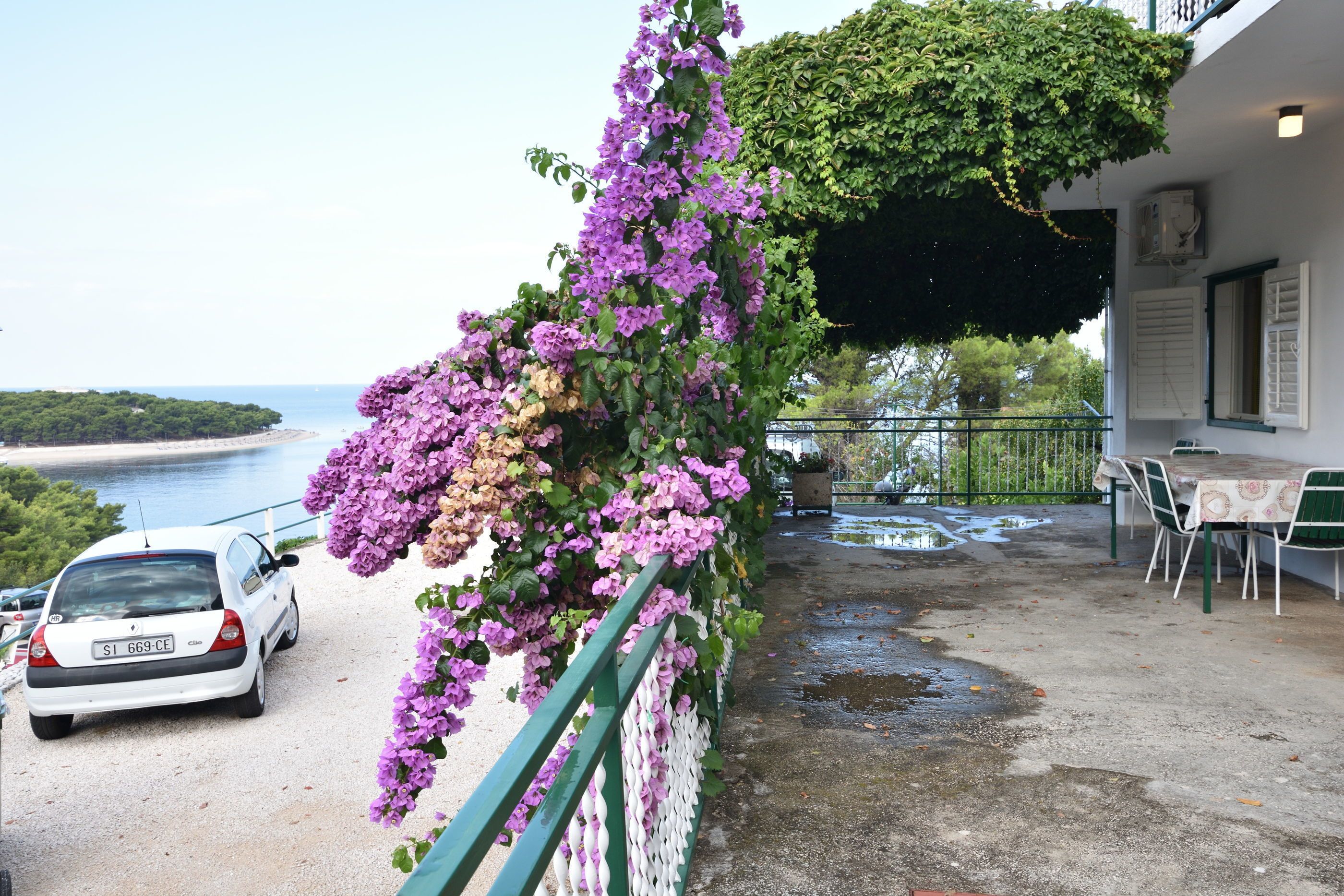 Ein Balkon mit einem parkenden Auto davor, einem Tisch und Stühlen sowie Blick auf das Meer.