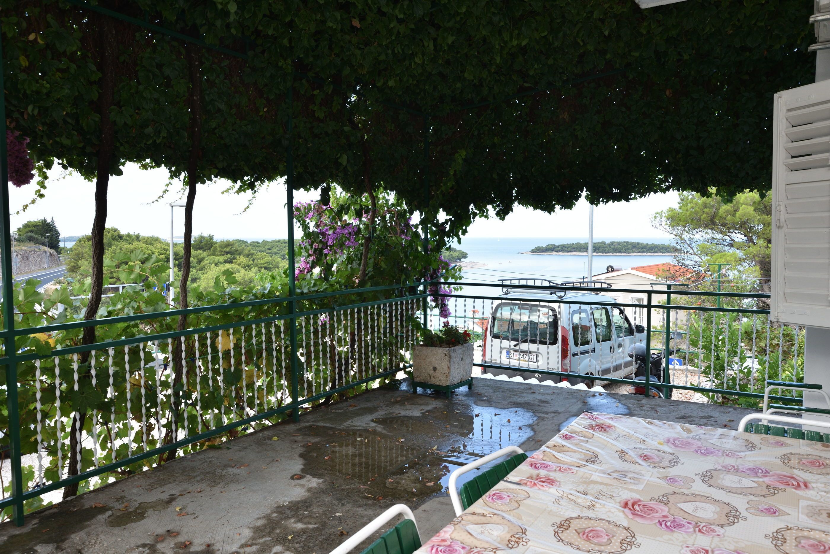 A balcony with a view of the sea, a parked van, and a table with a floral tablecloth.