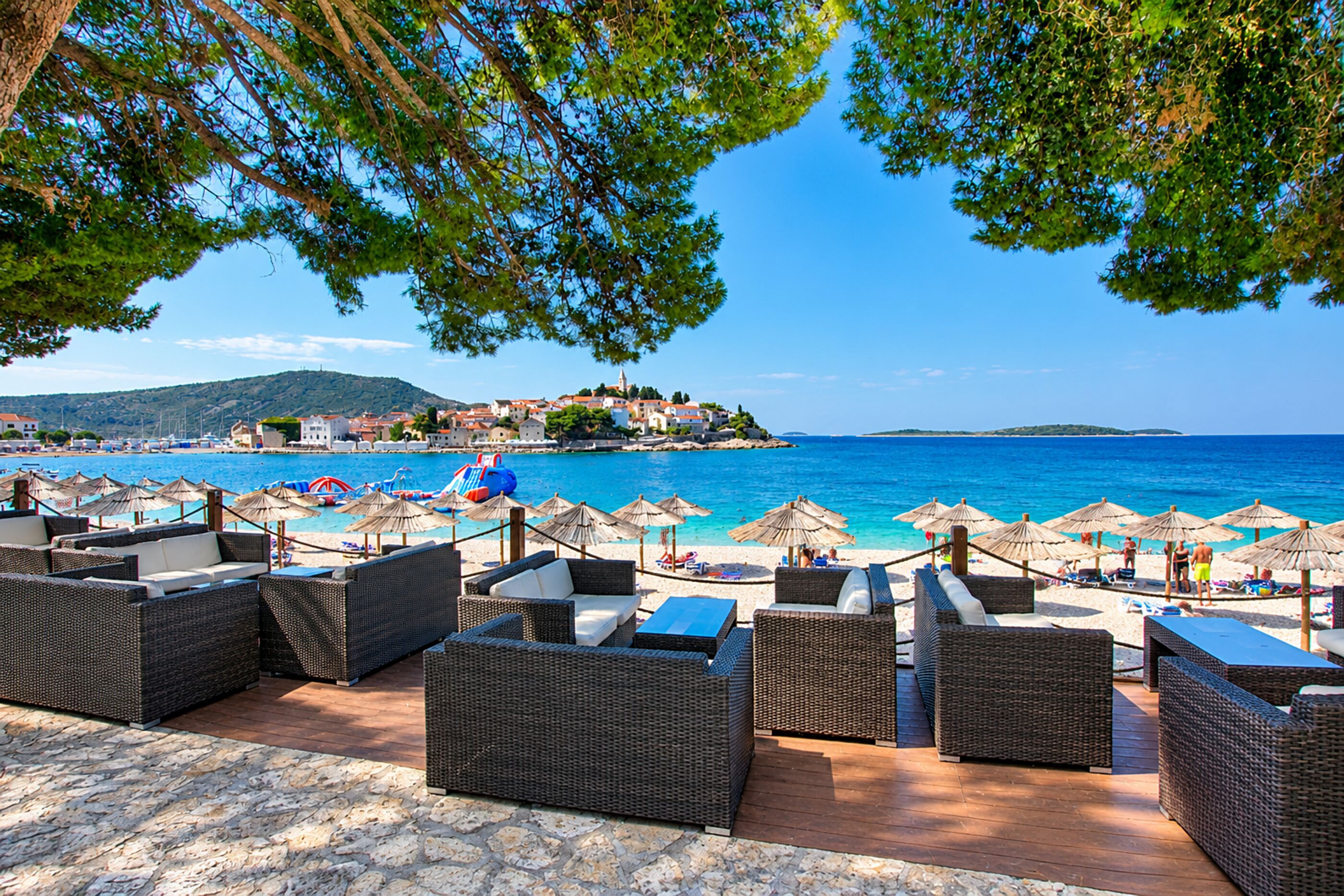 A beachside patio with wicker furniture and umbrellas, overlooking a clear blue sea and a coastal town.