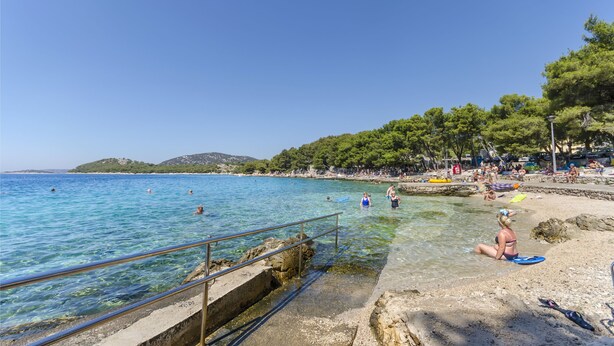 Ein Strand mit klarem, blauem Wasser, Menschen beim Schwimmen und Sonnenbaden sowie einer felsigen Küste.
