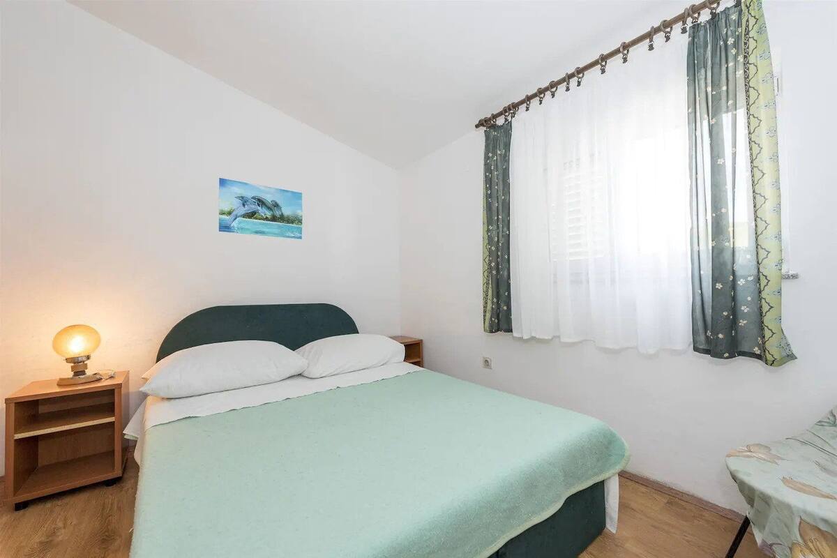 Un dormitori amb un llit, una nitòria amb una làmpada, una finestra amb cortines i una fotografia a la paret.