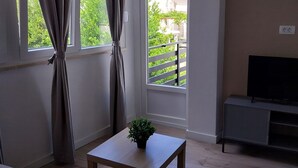 Ein Wohnzimmer mit einer Couch, einem Couchtisch mit Pflanze, einem Fernseher auf einem Schrank und einem Balkon mit Geländer.