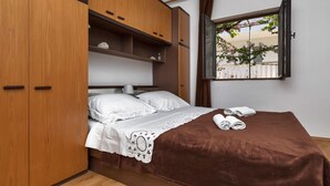 A bedroom with a bed, a brown blanket, two rolled towels, a window with a view of greenery, and wooden wardrobe with multiple compartments. - House Sea Rose (Zastrazisce)