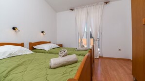 Ein Schlafzimmer mit zwei Betten, grüner Bettwäsche, Holz-Kopfteilen, weißen Wänden und einem Fenster mit durchsichtigen Vorhängen.