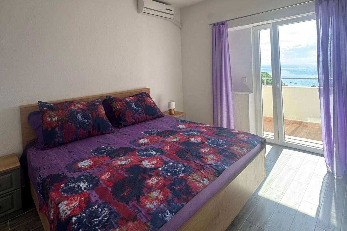 A bedroom with a bed, a ceiling fan, an air conditioning unit, and a view of the sea through a sliding glass door.