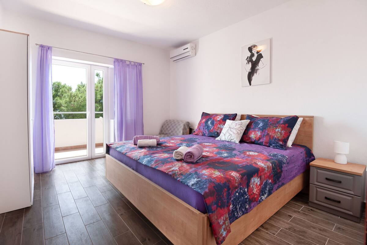 A bedroom with a bed, a nightstand, a window with curtains, and an air conditioning unit.