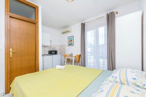Una habitación con cama, escritorio, silla, una kitchenette pequeña y ventana con cortinas.