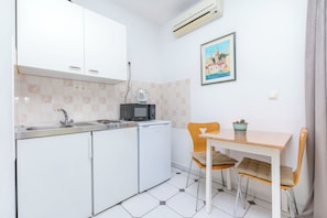 Cocina compacta con microondas, lavamanos y refrigerador pequeño. Cuenta con una mesa de comedor con dos sillas y una planta en maceta.