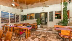 Un restaurant avec des tables et des chaises en bois, un sol carrelé en pierre et une vue sur le paysage grâce à de grandes fenêtres.