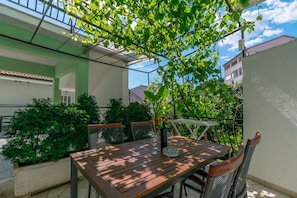 An outdoor patio with a wooden table and chairs, surrounded by greenery and a clear sky.