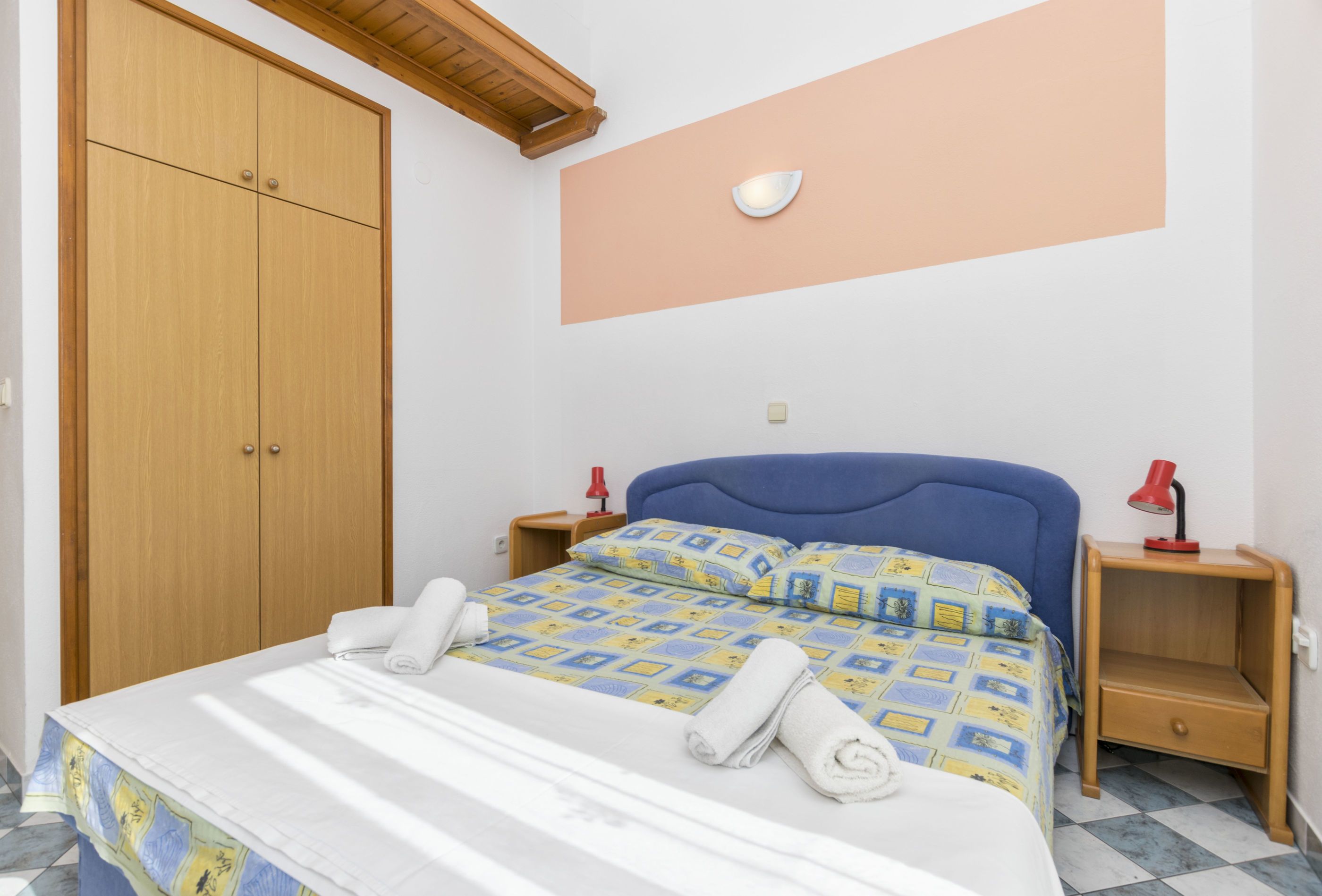 Een slaapkamer met een bed, een kledingkast, een nachtkastje met een lamp en een wandlamp.