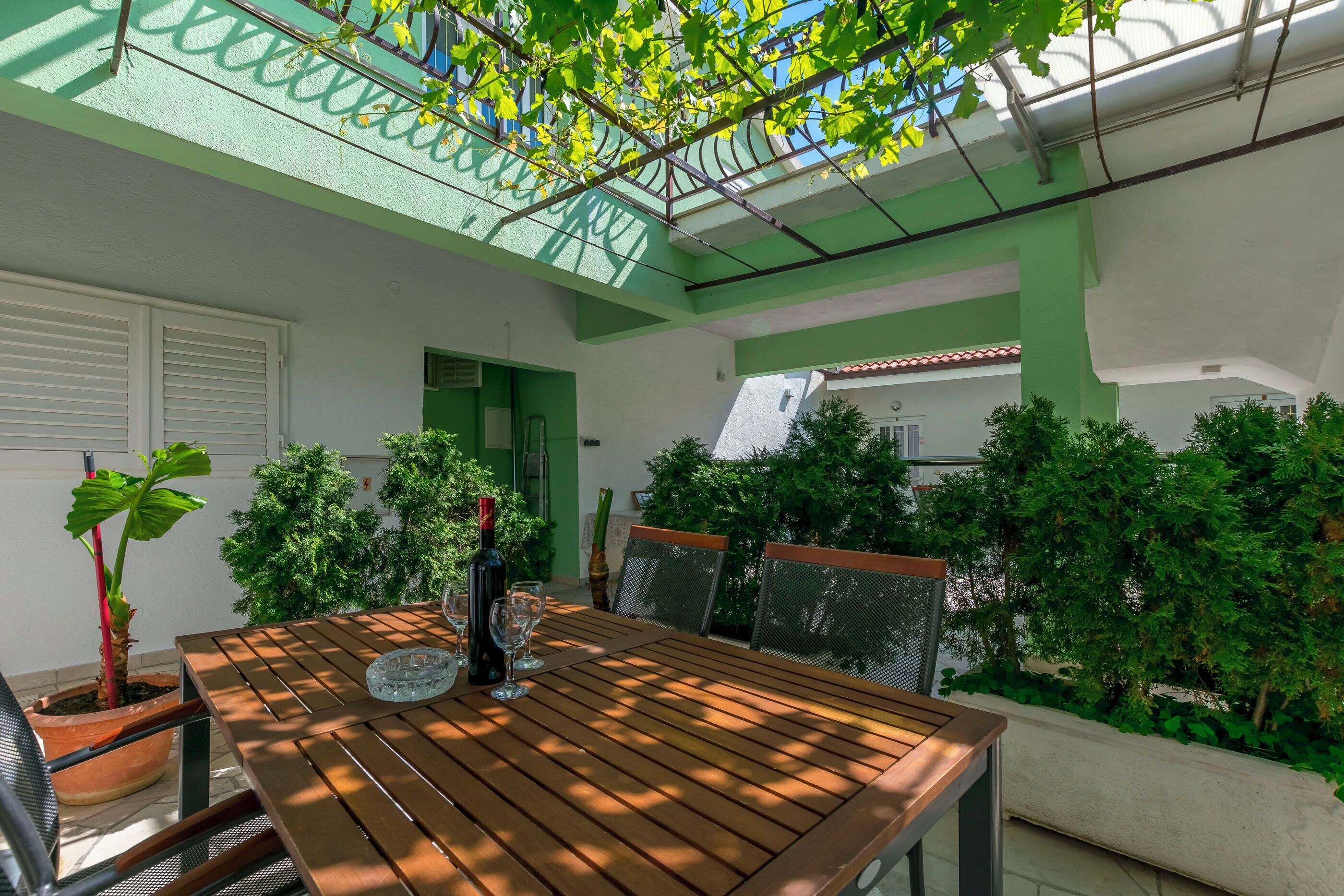 Une table en bois, entourée de chaises, avec une bouteille de vin et des verres, le tout agrémenté de plantes en pot et de verdure.