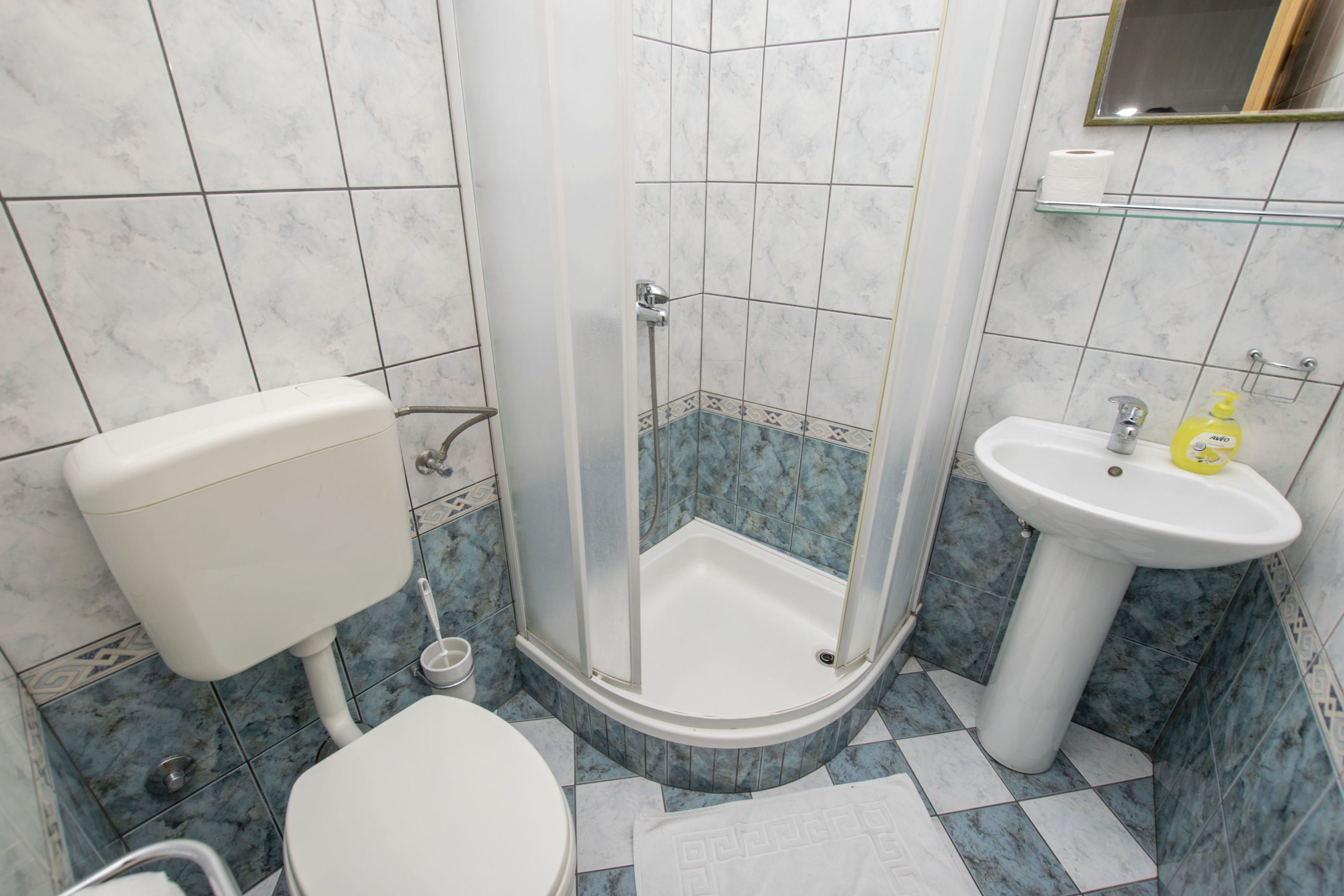 Egy fürdőszoba, amelyben található egy WC, egy üvegajtós zuhanyzó és egy mosdókagyló, felette pedig egy tükör.