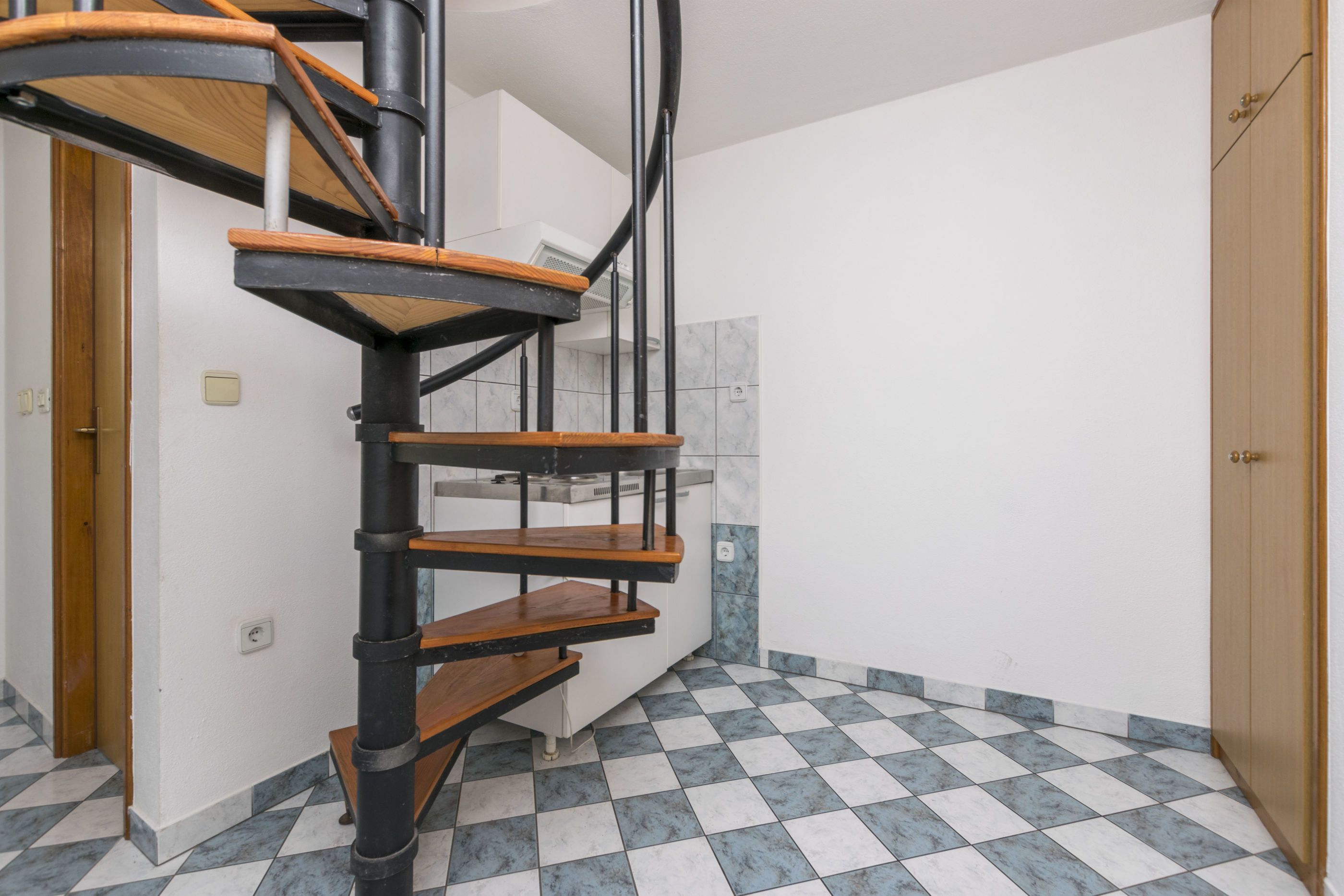 Una escalera de caracol con escalones de madera y barandillas de metal negro, que conduce a un rellano con pared blanca y suelo de baldosas.